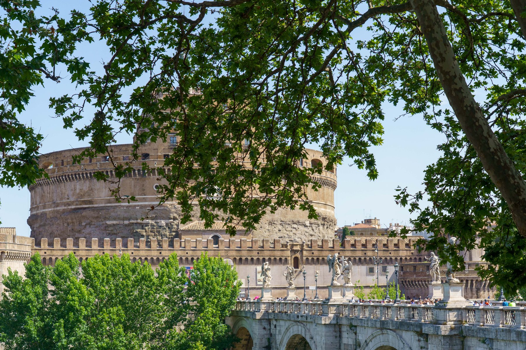 Castel Sant'Angelo seen through trees, with angel statues lining the bridge below