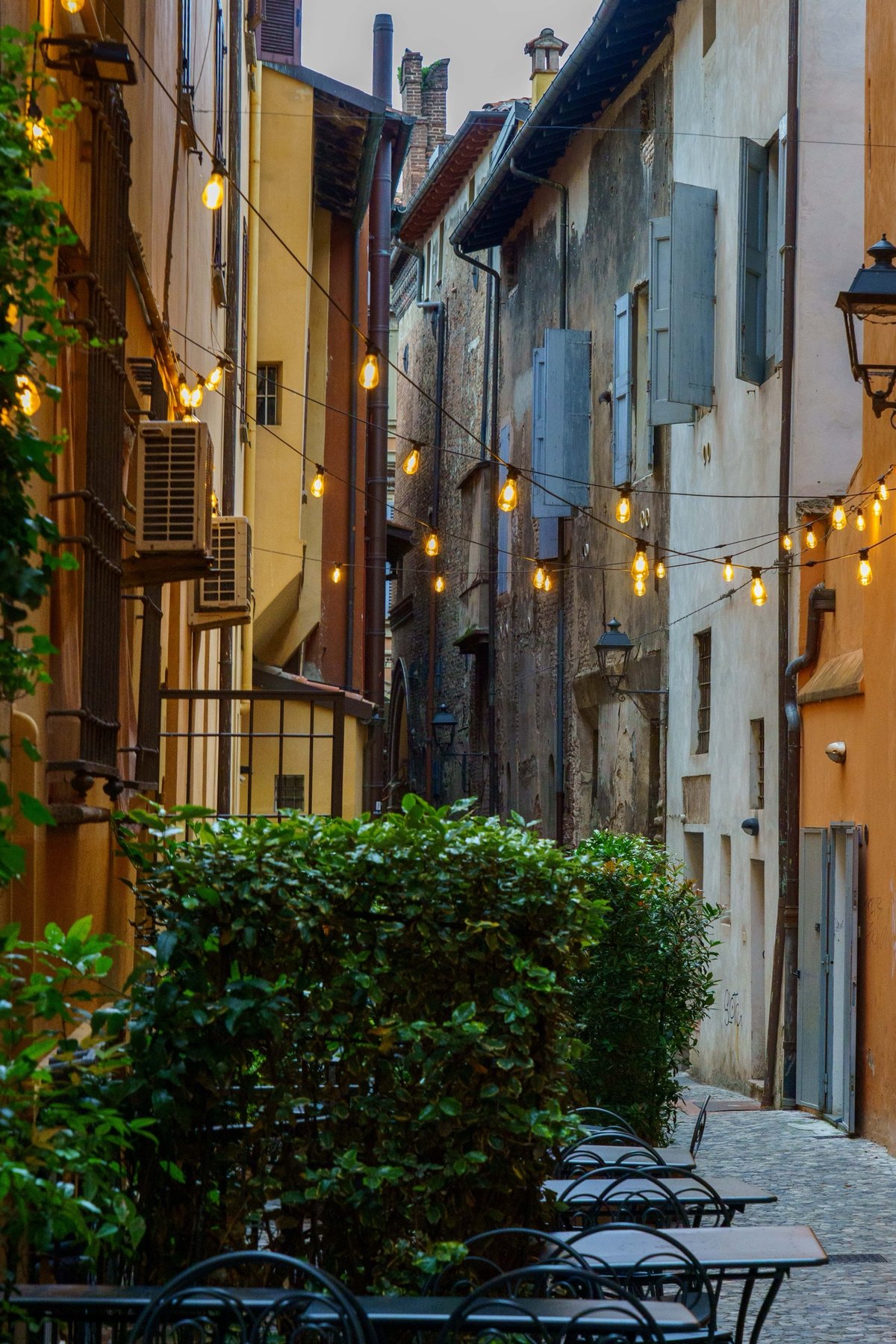 A quiet alley in Bologna strung with lights and lined with empty cafe tables at dusk