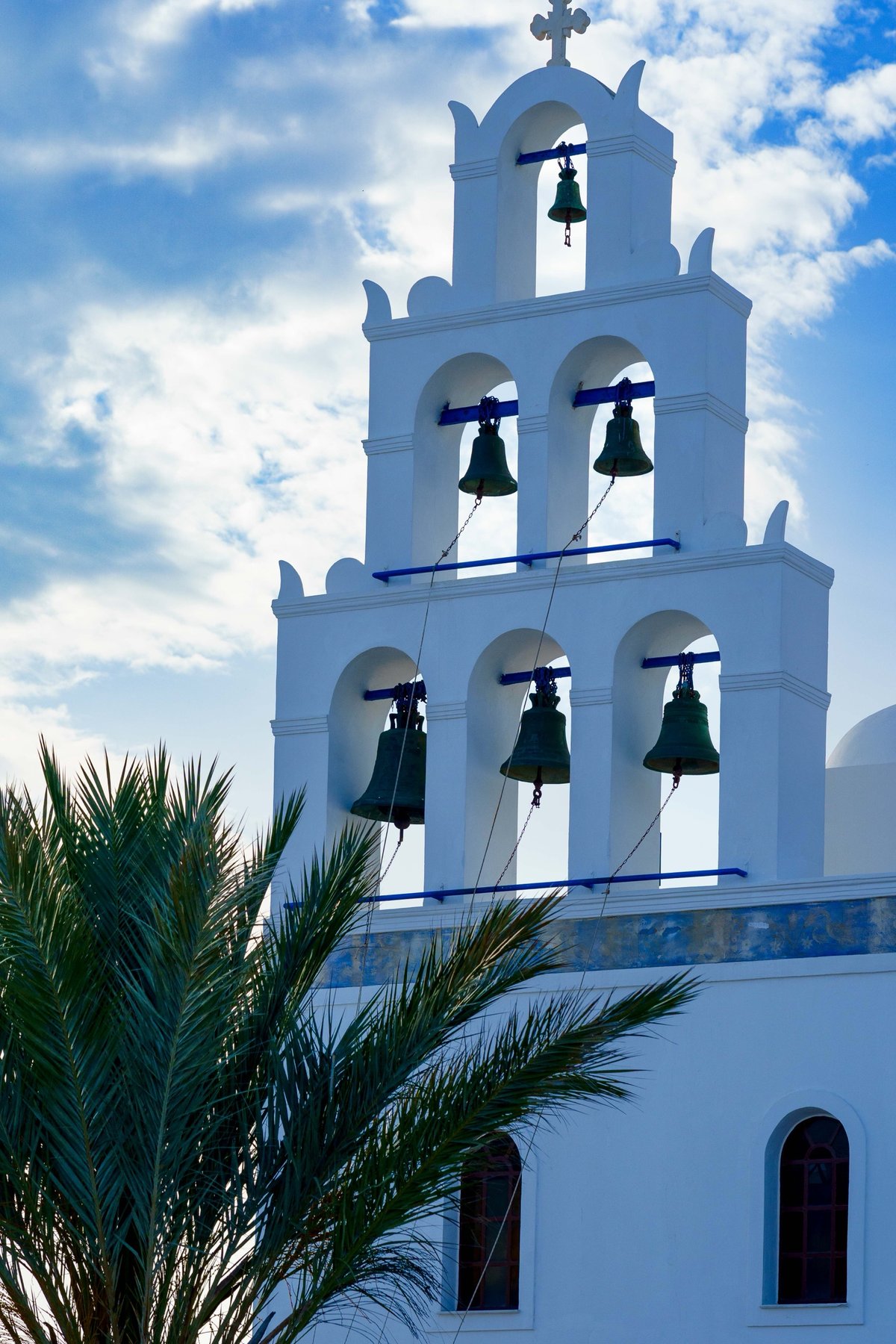 A whitewashed bell tower with its bells, framed by a palm tree in Santorini