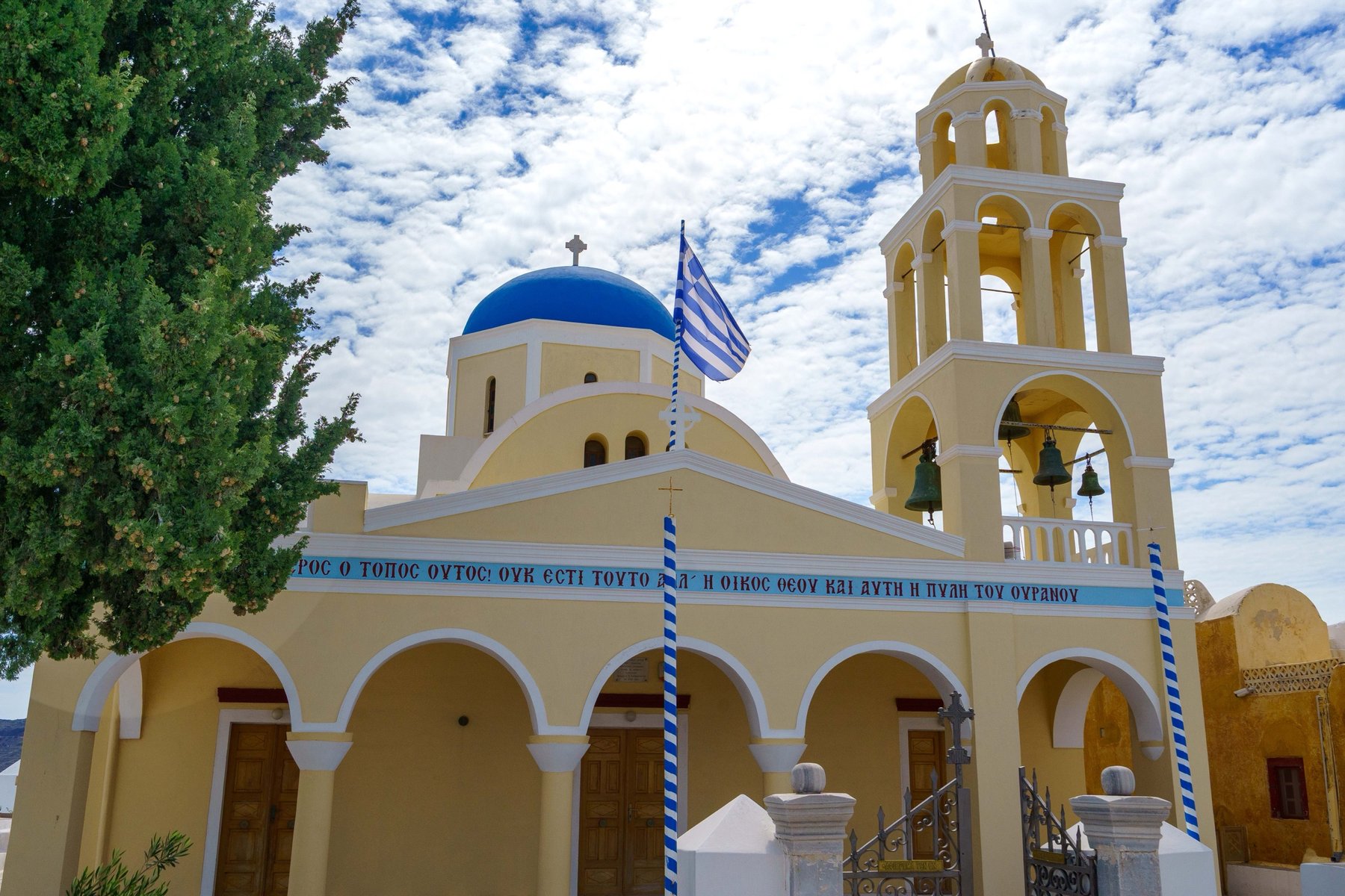 A yellow church with a blue dome and bell tower and a Greek inscription in Santorini