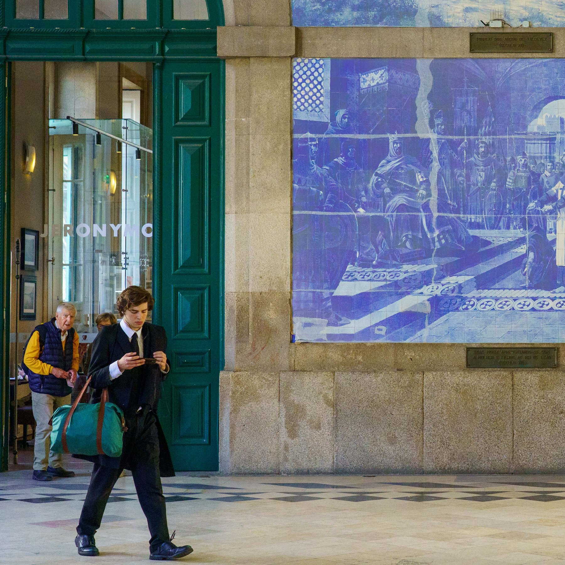 A young man absorbed in his phone walks past an older man at São Bento Station, its walls covered in historic azulejo tile murals