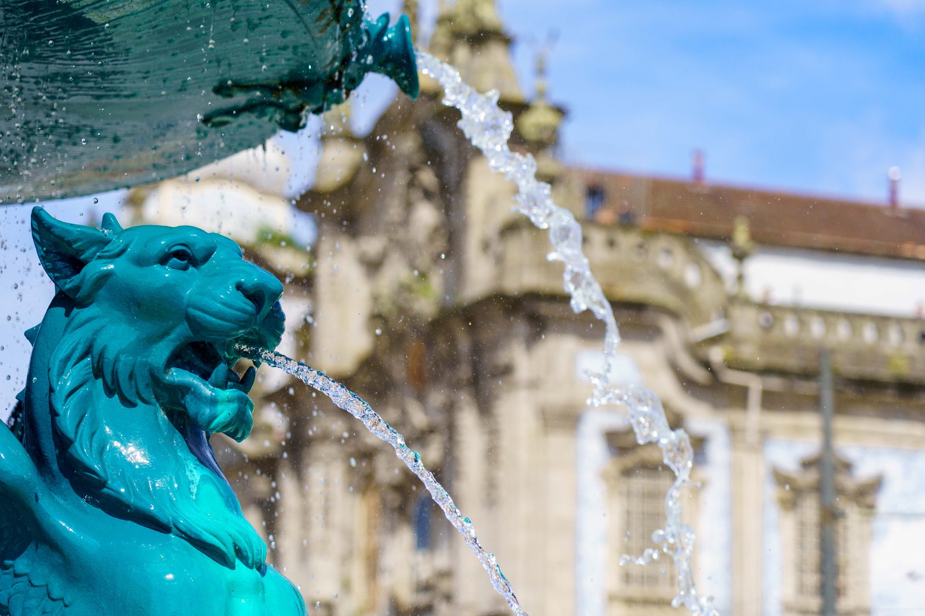 A teal lion-head fountain spout with the University of Porto building blurred in the background